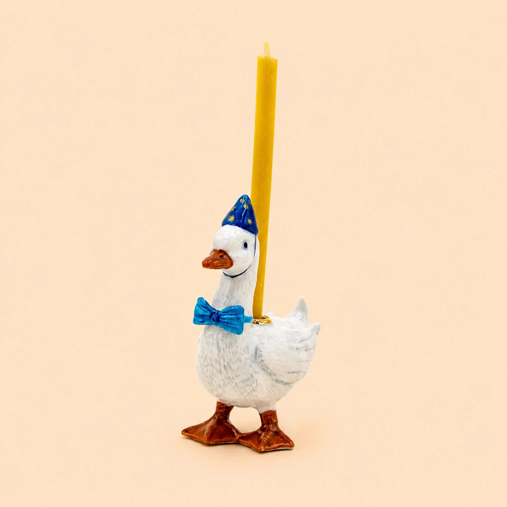 Introducing the Blue Goose Cake Topper – Final Sale: this handcrafted ceramic figurine features a goose wearing a blue bow tie and party hat, carrying a yellow candle on its back, beautifully photographed against a plain beige background.