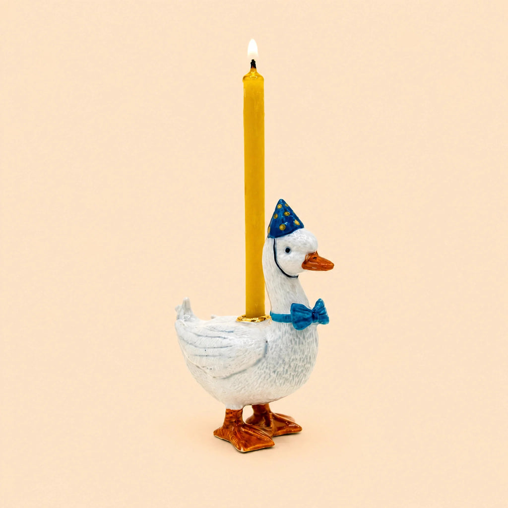 A white goose figurine, called the “Blue Goose Cake Topper – Final Sale,” wears a blue party hat and bow tie while carrying a lit yellow candle on its back, set against a plain beige background. This charming porcelain cake topper makes a delightful collectible keepsake for any celebration.