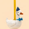 The Blue Goose Cake Topper—featuring a handcrafted white goose in a blue party hat with yellow dots, a matching blue bow tie, and carrying a yellow candle on its back—serves as an adorable birthday decoration against a plain light peach background.