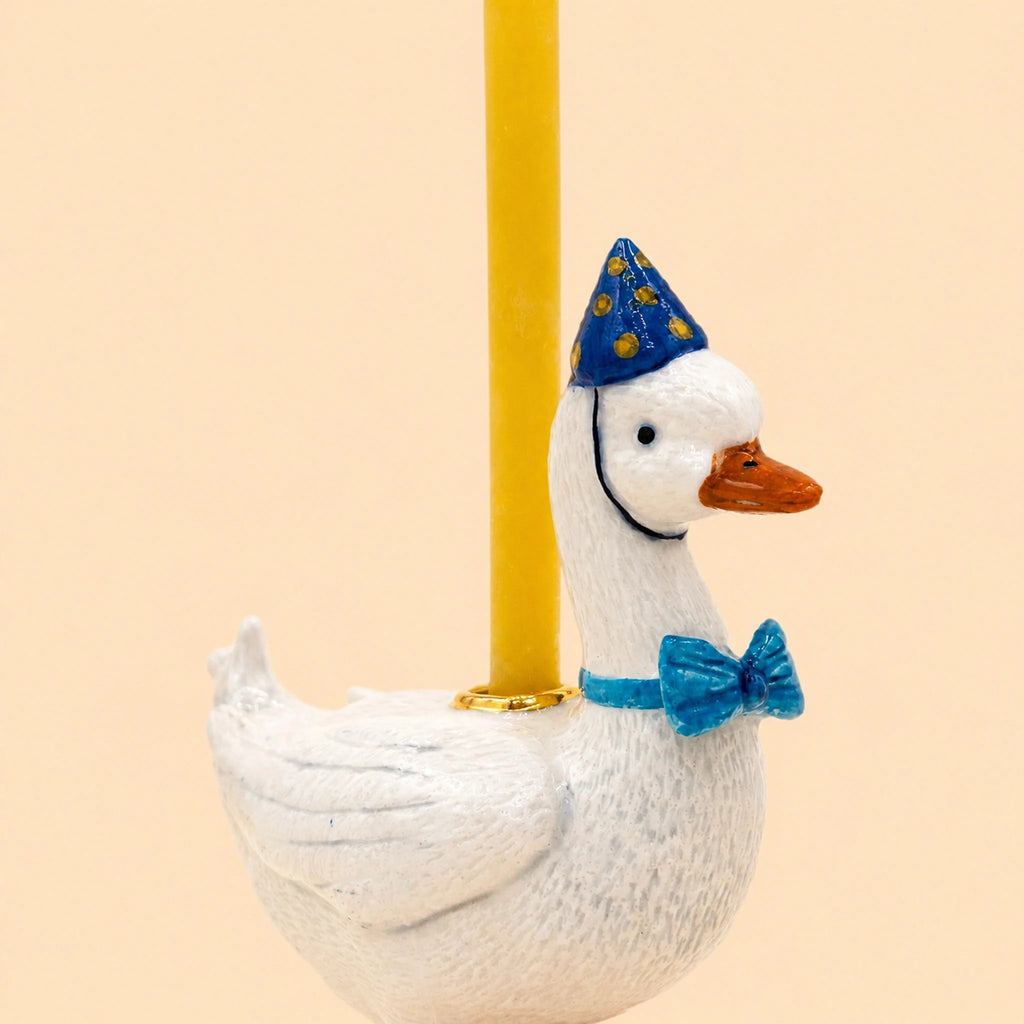 The Blue Goose Cake Topper—featuring a handcrafted white goose in a blue party hat with yellow dots, a matching blue bow tie, and carrying a yellow candle on its back—serves as an adorable birthday decoration against a plain light peach background.
