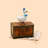 A charming Blue Goose Cake Topper, this ceramic duck is dressed in a blue party hat and bow tie, standing atop a small wooden chest placed on a beige star-patterned cloth, with a simple peach background behind it.