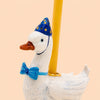 Introducing the Blue Goose Cake Topper - Final Sale: this handcrafted porcelain birthday decoration features a white goose figurine donning a blue party hat with yellow dots and a coordinating bow tie, balancing a yellow candle on its back. Ideal as a charming cake topper or collectible keepsake, it is showcased against a plain light peach background.