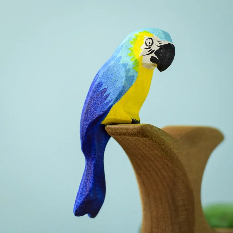 The Bumbu Wooden Blue Macaw Parrot wooden toy, painted in vibrant blue, yellow, and green hues, is perched on a wooden branch. The background is a plain light blue, showcasing the parrot's bright and colorful appearance. Montessori & Waldorf Inspired, it’s crafted from eco-friendly materials for guilt-free fun.