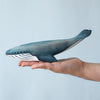 A person holds a Bumbu Handmade Wooden Blue Whale Family Set figurine, painted blue and white, against a light blue background—ideal for Montessori play or as a sustainable wood toy.