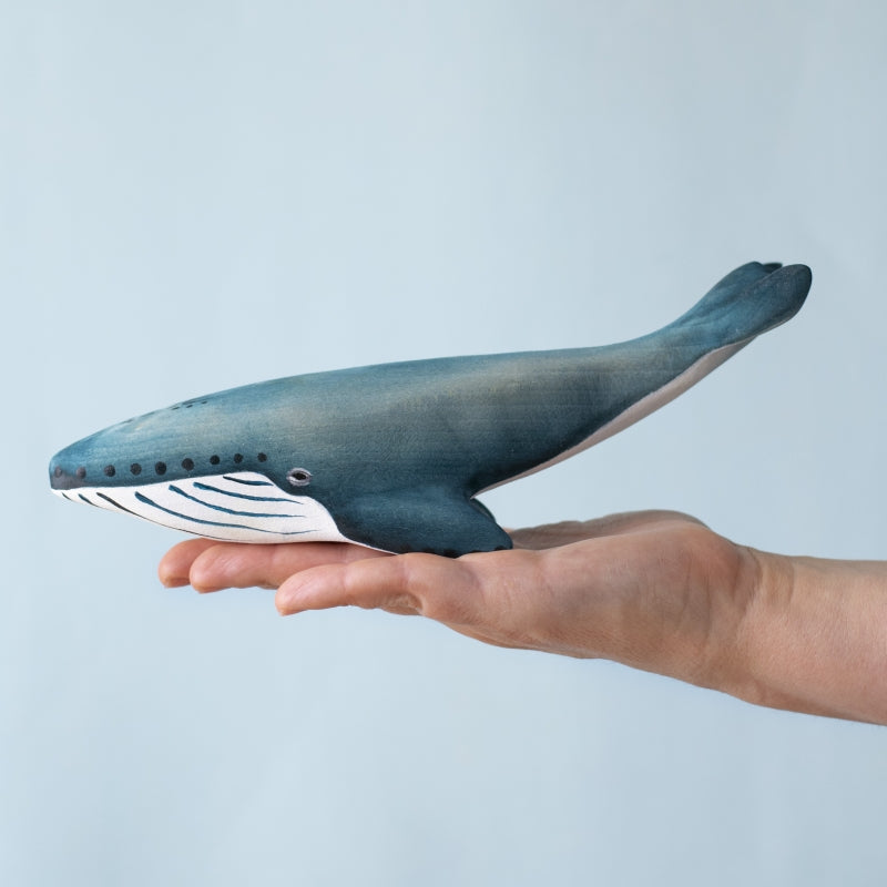 A person holds a Bumbu Handmade Wooden Blue Whale Family Set figurine, painted blue and white, against a light blue background—ideal for Montessori play or as a sustainable wood toy.
