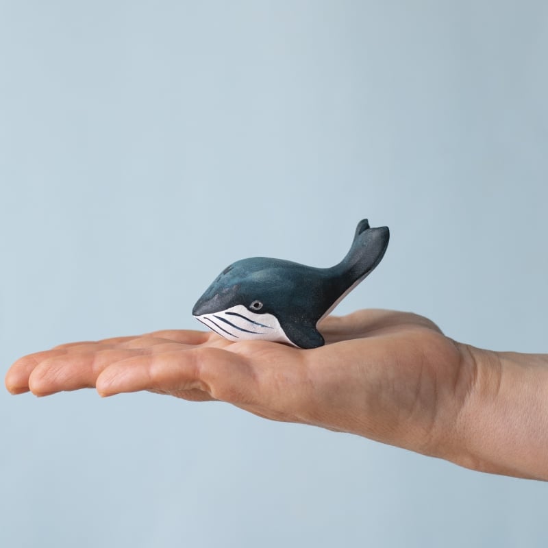 A hand holds a small black and white whale figurine from the Bumbu Handmade Wooden Blue Whale Family Set, ideal for Montessori play, against a light blue background.