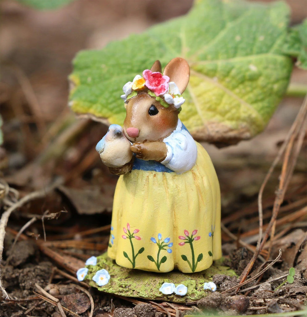 The Wee Forest Folk Tiny Collectible - Bluebird of Happiness (Not a Toy) features a handmade mouse in a yellow floral dress with a flower crown, gently holding a bird, standing on soil with green leaves and small painted flowers.