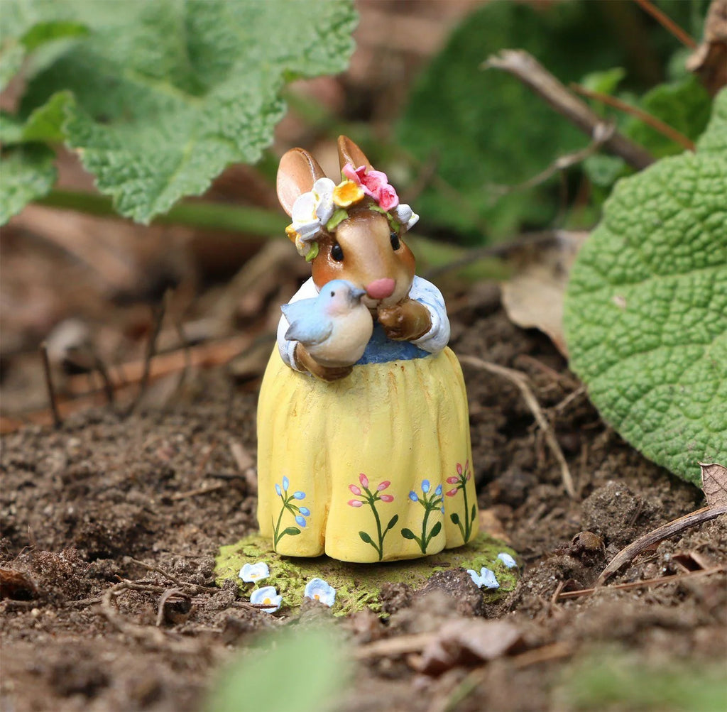 The Wee Forest Folk Tiny Collectible - Bluebird of Happiness features a rabbit in a flower crown and yellow skirt, holding a blue and white bird, standing on soil—a charming hand-sculpted miniature for collectors.
