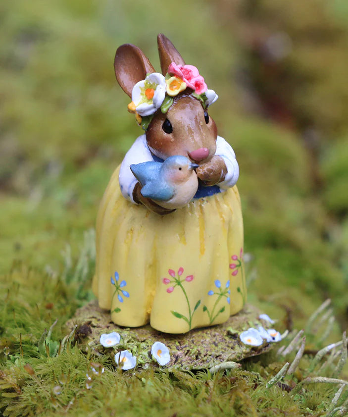 Hand sculpted, the Wee Forest Folk Tiny Collectible - Bluebird of Happiness features a brown bunny in a yellow floral dress and flower crown, standing on moss with a blue bird, surrounded by white and yellow flowers.