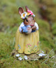 Hand sculpted, the Wee Forest Folk Tiny Collectible - Bluebird of Happiness features a brown bunny in a yellow floral dress and flower crown, standing on moss with a blue bird, surrounded by white and yellow flowers.
