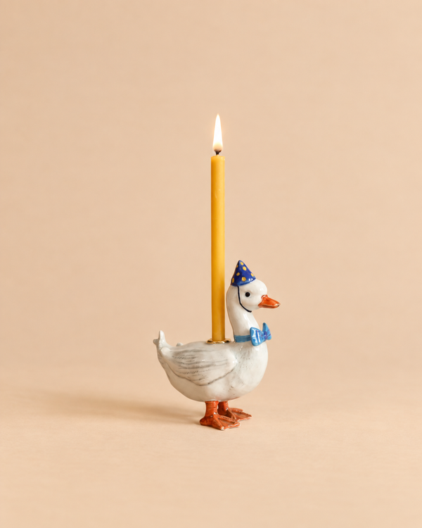 Introducing the Blue Goose Cake Topper – Final Sale: a handcrafted collectible featuring a white ceramic goose wearing a blue party hat and bow tie, with a lit yellow candle standing upright on its back, beautifully set against a beige background.