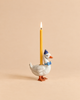 Introducing the Blue Goose Cake Topper – Final Sale: a handcrafted collectible featuring a white ceramic goose wearing a blue party hat and bow tie, with a lit yellow candle standing upright on its back, beautifully set against a beige background.