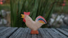 A small handmade Holzwald Bremer Rooster wooden figurine with a red comb and a multi-colored tail, standing on a wooden surface.