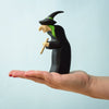 A hand with red nail polish holds the Bumbu Handmade Wooden Brewing Witch and Cauldron Set, featuring a witch with a pointed hat, green hair, long nose, and broom against a light blue background.