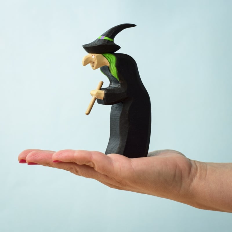 A hand with red nail polish holds the Bumbu Handmade Wooden Brewing Witch and Cauldron Set, featuring a witch with a pointed hat, green hair, long nose, and broom against a light blue background.