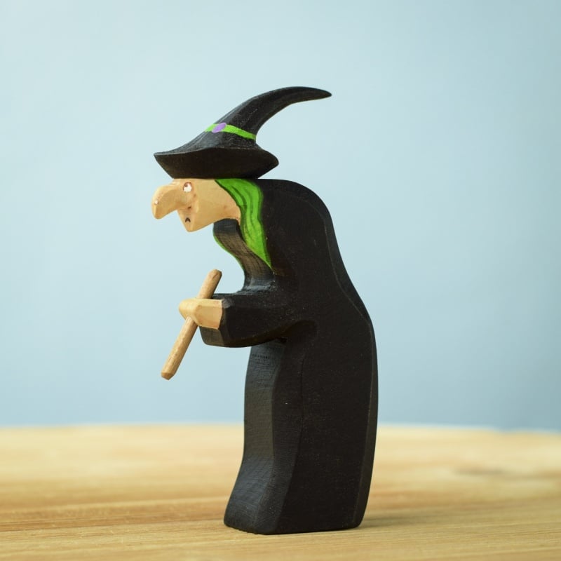 The Bumbu Handmade Wooden Brewing Witch and Cauldron Set features a witch with a pointed hat, green hair, hooked nose, and black cloak holding a brown staff. This Montessori Waldorf toy is shown on wood with a blue background.