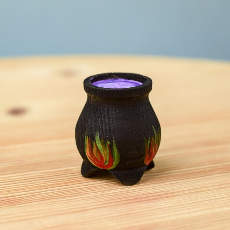 The Bumbu Handmade Wooden Brewing Witch and Cauldron Set features a small black cauldron with red and green flames, filled with purple material on a light wooden surface—a magical toy perfect for imaginative play.