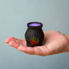 A hand holds the Bumbu Handmade Wooden Brewing Witch and Cauldron Set—a small black cauldron with red and green flames, filled with purple liquid—against a plain light blue background.