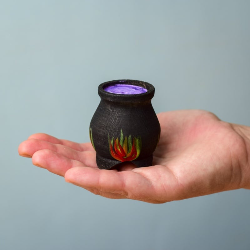 A hand holds the Bumbu Handmade Wooden Brewing Witch and Cauldron Set—a small black cauldron with red and green flames, filled with purple liquid—against a plain light blue background.