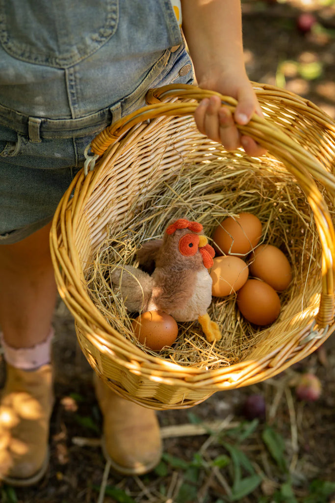 A person wearing denim overalls and boots is holding a wicker basket filled with hay, six brown eggs, a small plush chicken toy, and a Bubba Rooster Rattle. The background is a blurred outdoor setting with grass and sunlight filtering through.