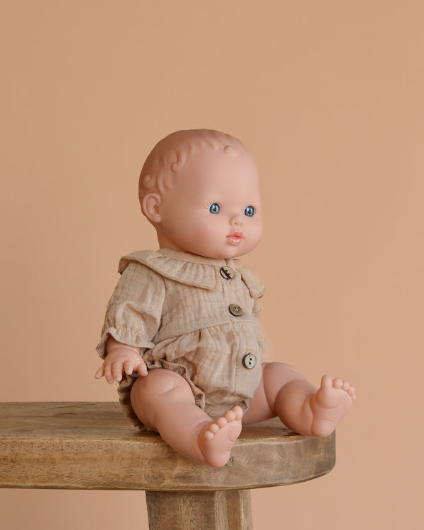 The Minikane Baby Doll (13") - Rosalie with Clothing, featuring blue eyes and a beige buttoned romper, sits on a wooden stool against a plain beige background. This anatomically correct doll is beautifully crafted in Spain.