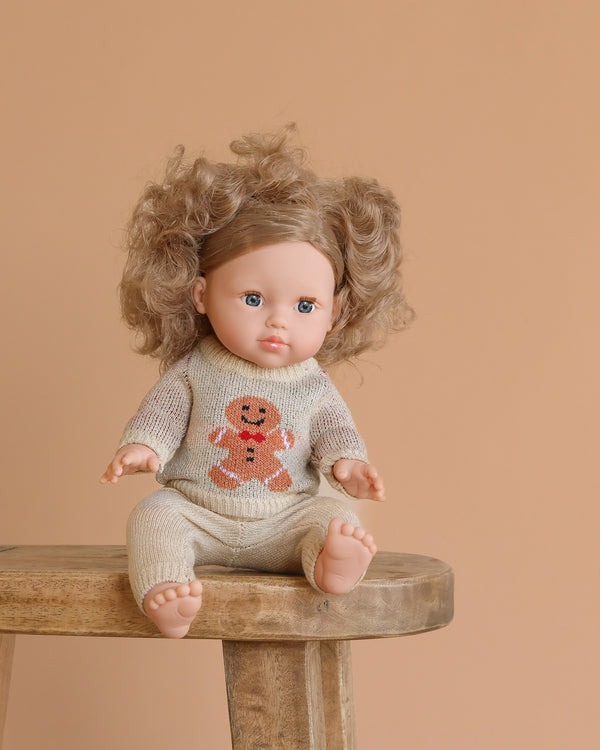 The Minikane Baby Doll (13") - Lola with Clothing, featuring curly hair and blue eyes, sits on a wooden stool in a beige knit gingerbread sweater. This anatomically correct doll is made in Spain and displayed against a peach background.