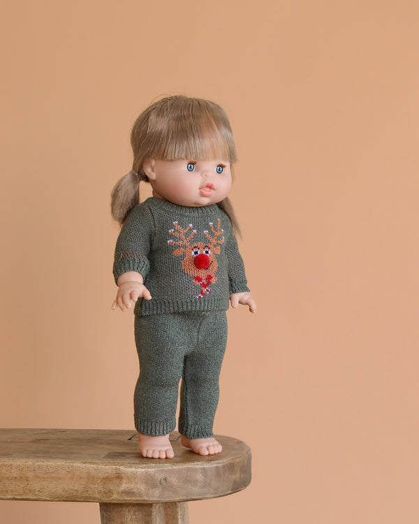 The Minikane Standing Doll (14") - Yzé with Clothing, featuring light brown hair and bangs, stands barefoot on a wooden stool in a green knit outfit with a red reindeer against a peach background. This anatomically correct doll is made in Spain.