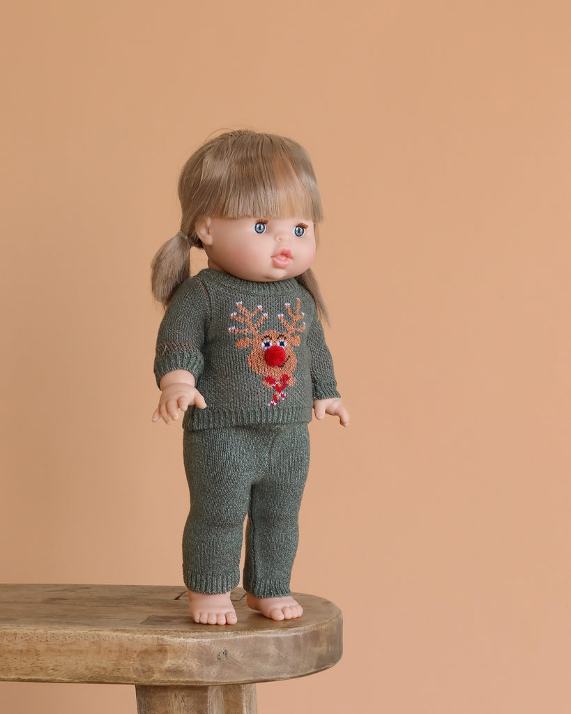 The Minikane Standing Doll (14") - Yzé with Clothing, featuring light brown hair and bangs, stands barefoot on a wooden stool in a green knit outfit with a red reindeer against a peach background. This anatomically correct doll is made in Spain.