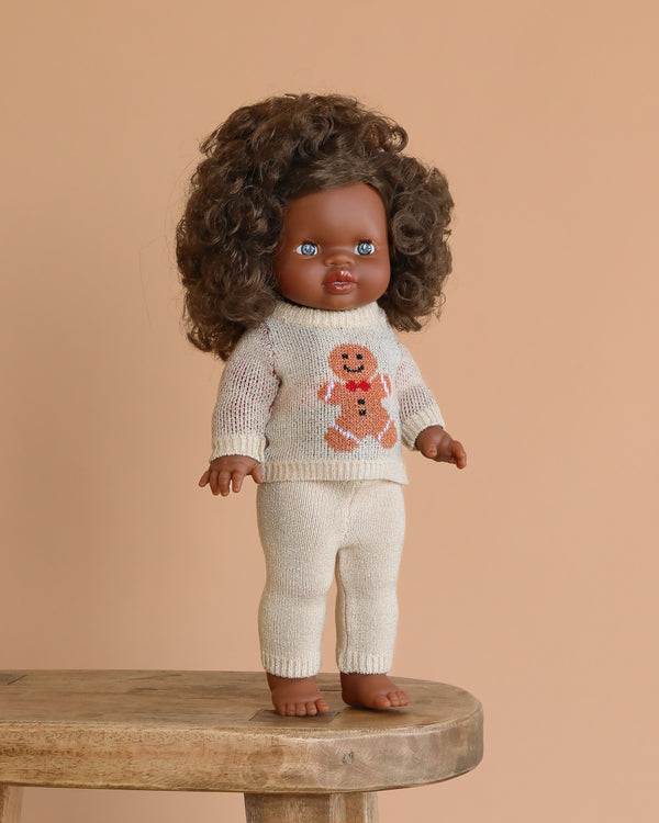 The Minikane Standing Doll (14") - Charlie with Clothing features a dark-skinned baby doll with curly brown hair in a cream knit sweater with a gingerbread man design and matching pants, posed on a wooden stool against a beige background.