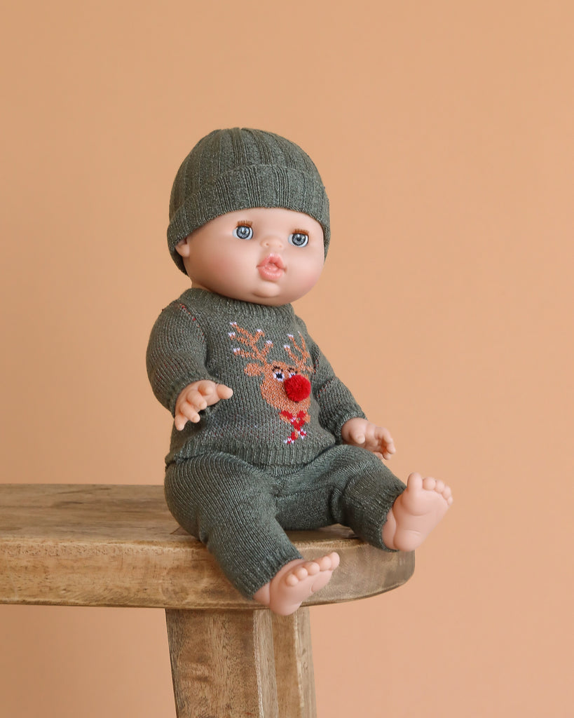 The Minikane Doll with Sleeping Eyes (13") – Lily with Clothing, made in Spain, sits on a wooden stool wearing a green knit hat and reindeer outfit against a peach-colored background.
