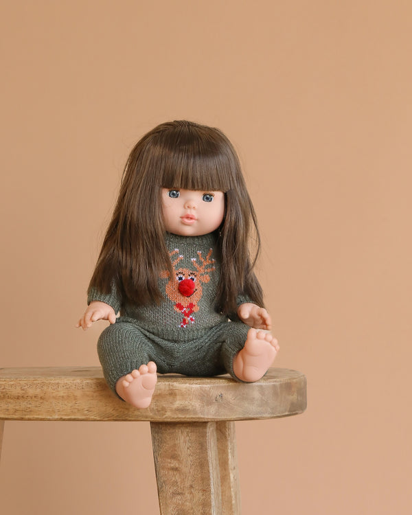The Minikane Baby Doll (13") - Cherry with Clothing, in a green knit outfit with a red reindeer, sits on a wooden stool against a plain beige background. She has long brown hair and bangs.