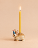 The Bunny Rabbit Cake Topper - Final Sale features a ceramic rabbit figurine wearing a blue party hat with a red tip, holding a lit yellow candle, reminiscent of a handcrafted collectible and set against a neutral beige background.
