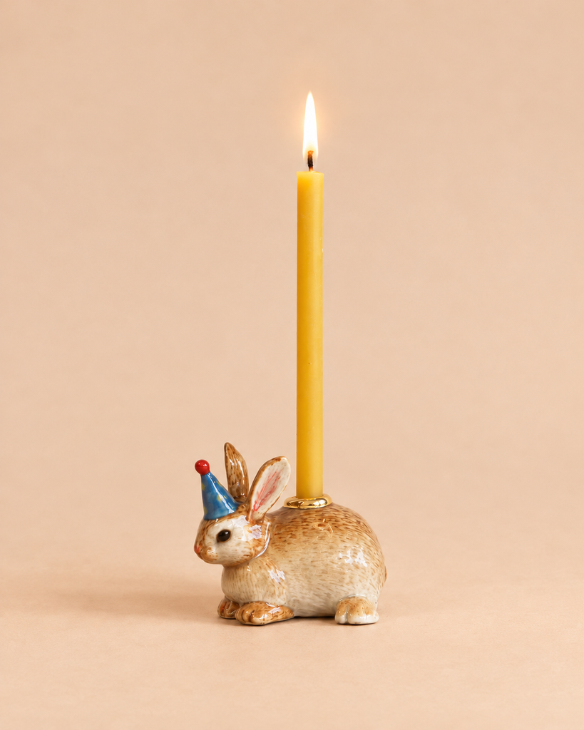 The Bunny Rabbit Cake Topper - Final Sale features a ceramic rabbit figurine wearing a blue party hat with a red tip, holding a lit yellow candle, reminiscent of a handcrafted collectible and set against a neutral beige background.