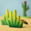 A close-up photograph of two colorful toy cacti with bright green spikes, part of the Bumbu Wooden Bushes Set crafted from sustainably sourced wood. In the blurred background, there is a larger cactus with red flowers on top. The setting appears to be on a beige surface with a light blue backdrop.