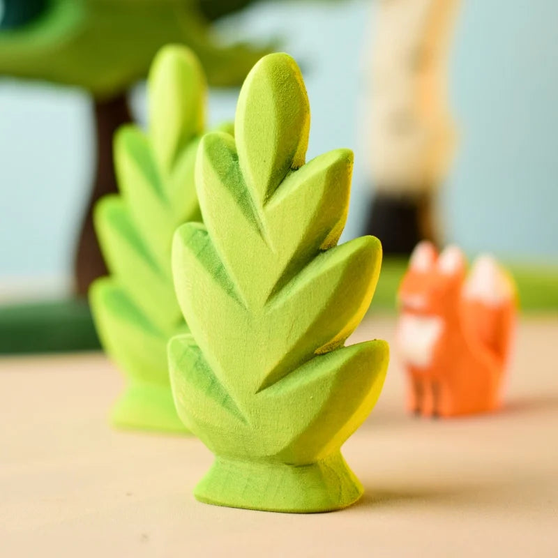 Close-up of two bright green, leaf-shaped wooden toys from the Bumbu Wooden Bushes Set with an orange, fox-shaped toy in the background. The focus is on the eco-friendly toys, with the orange fox slightly blurred. Inspired by Montessori & Waldorf principles, this set is perfect for imaginative indoor play.