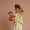A cheerful young child with curly hair smiles and holds an Olli Ella | Dinkum Doll - Button, wearing a yellow top and striped pants against a soft beige background.