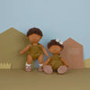 Two posable Olli Ella Dinkum Dolls with dark curly hair, dressed in green overalls, sitting against a backdrop of simple geometric shapes resembling houses, in shades of brown and beige.