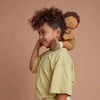 A young boy with curly hair smiles gently while holding an Olli Ella Dinkum Doll - Button on his shoulder, both wearing matching green shirts against a soft peach background.
