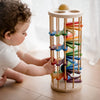 A toddler with curly hair wearing a white outfit is playing with the Pound A Ball Tower, a vertical wooden toy made from non-toxic materials. This cylindrical toy features a rainbow track with multiple colorful ramps designed to develop motor skills as the ball rolls down from top to bottom. The child is seen pushing one of the ramps.