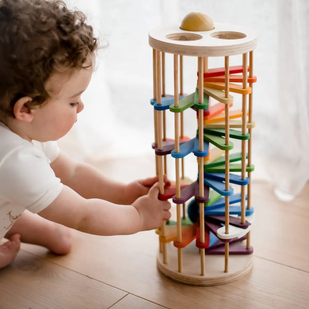 A toddler with curly hair wearing a white outfit is playing with the Pound A Ball Tower, a vertical wooden toy made from non-toxic materials. This cylindrical toy features a rainbow track with multiple colorful ramps designed to develop motor skills as the ball rolls down from top to bottom. The child is seen pushing one of the ramps.