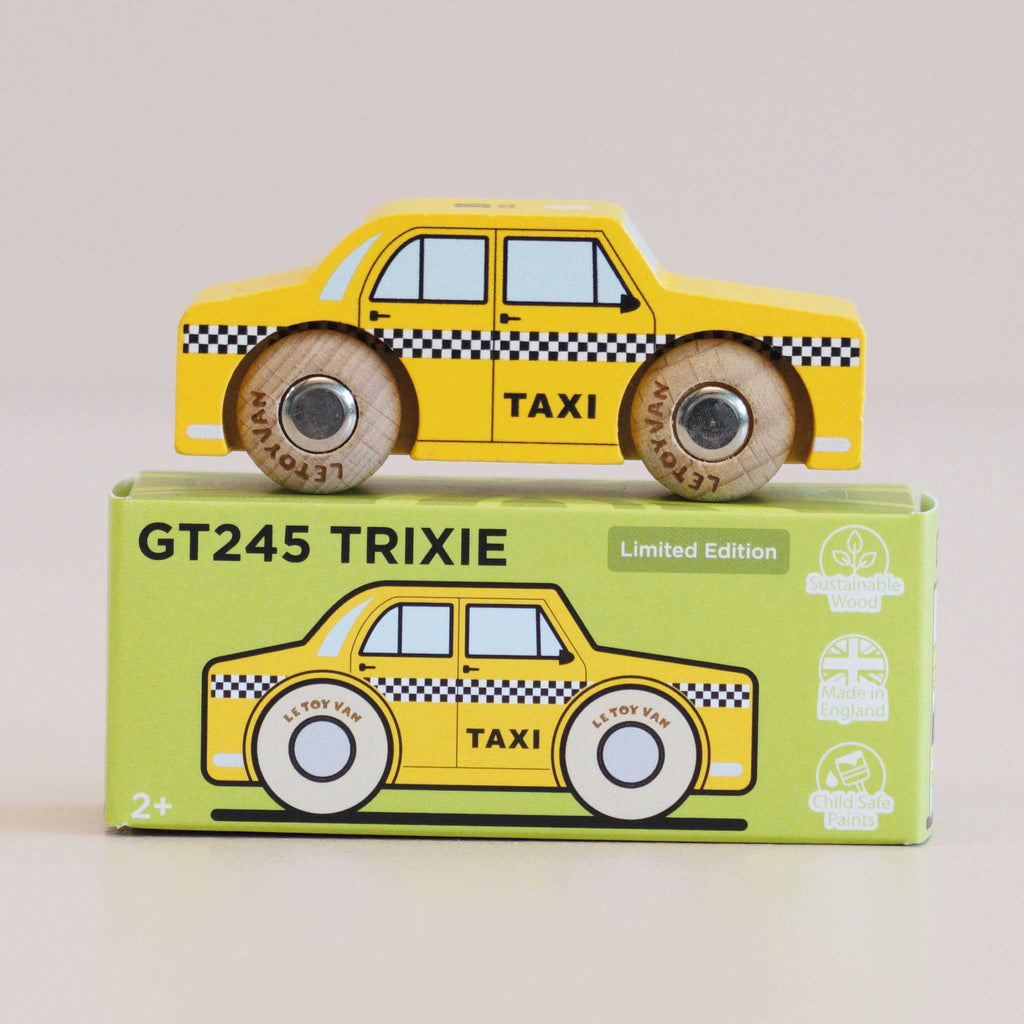 A yellow wooden taxi toy, named No. 39 GT245 Trixie and made in England, sits atop its green box packaging, which displays an illustration of the New York taxi along with the text “No. 39 GT245 Trixie | Made in England” and “Limited Edition.” Black and white checks decorate the side of the toy, making it ideal for imaginative play.