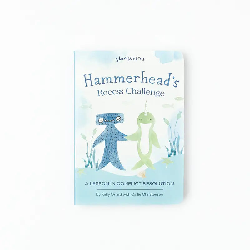 The image is of the book cover for "Slumberkins Hammerhead Kin + Lesson Book - Conflict Resolution" by Kelly Oriard and Callie Christensen. The cover illustration features a blue hammerhead shark and a green narwhal standing side by side, holding hands, emphasizing social skills and emotional regulation.