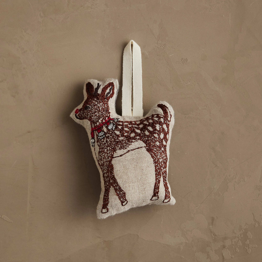The Coral & Tusk Rudolph Ornament is a small, soft reindeer shaped like a fawn with a red collar and white spots, hanging by a cream loop against a textured beige wall.