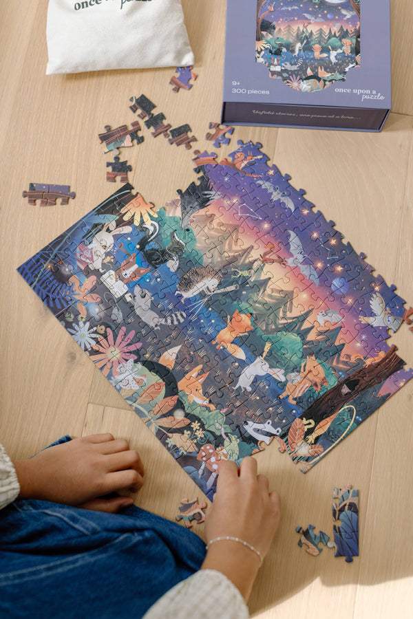 A person assembles the Mischief Under the Moonlight 300 Piece Puzzle with animal and forest designs. Matte pieces are scattered on a light wood surface next to the box, while a fabric pouch peeks out at the top left.
