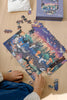 A person assembles the Mischief Under the Moonlight 300 Piece Puzzle with animal and forest designs. Matte pieces are scattered on a light wood surface next to the box, while a fabric pouch peeks out at the top left.