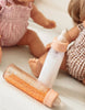 Two dolls wearing patterned outfits are positioned on a white surface, resembling The Essentials from the Tiny Tummies Pink Gift Box by Tiny Harlow. In front of them are two baby bottles with beige caps—one filled with orange liquid and the other with white. This scene encapsulates a playful, child-like essence perfect for tiny tummies.