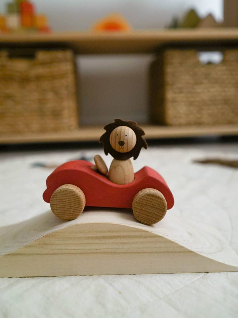 A Sabo Concept First Journey wooden toy lion rides in a red car on a wooden ramp, sparking imagination and open-ended play, with woven baskets and toys softly blurred in the background.