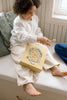 A young child with curly hair, dressed in a white shirt and cream pants, smiles while sitting on a window seat with the Honeycomb Cafe 200 Piece Puzzle and a golden box on their lap.