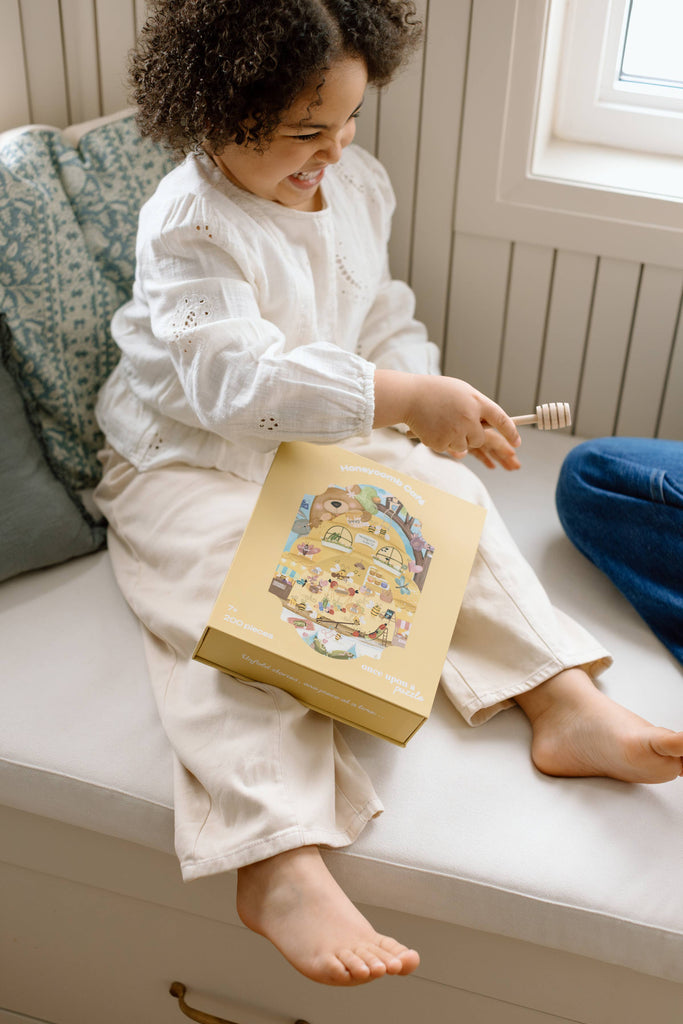 A young child with curly hair, dressed in a white shirt and cream pants, smiles while sitting on a window seat with the Honeycomb Cafe 200 Piece Puzzle and a golden box on their lap.