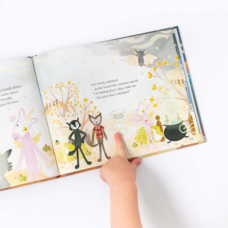 A child's hand points to a colorful illustrated page in the Slumberkins "Halloween Fright" Hardcover Book featuring whimsical animals and magical creatures under a starry, spooky season sky.